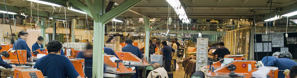 Pracovníci v továrně sedí u stolů a pracují na různých úkolech. Továrna je plná strojů a vybavení, s několika pracovníky v modrých uniformách soustředěných na svou práci.